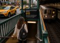 Une voyageuse descend l'escalier vert d'une station de métro de New York, un sac à dos sur l'épaule, tandis qu'une rame de la ligne R s'approche du quai et que des taxis jaunes circulent en surface.