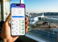 Smartphone affichant une application de comparaison de vols pas chers dans un terminal d'aéroport avec des avions en arrière-plan.