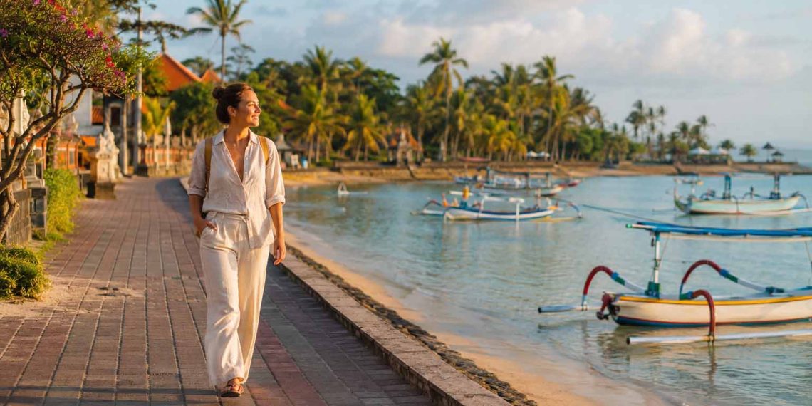 Bali sans scooter : est-ce vraiment possible ? 1 Une jeune femme marche à pied sur la promenade pavée bordée de palmiers le long de la plage de Sanur à Bali, illustrant les alternatives de transport sans scooter.