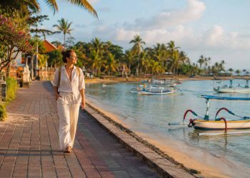 Bali sans scooter : est-ce vraiment possible ? 2 Une jeune femme marche à pied sur la promenade pavée bordée de palmiers le long de la plage de Sanur à Bali, illustrant les alternatives de transport sans scooter.