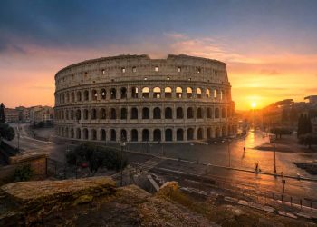 Vue panoramique du Colisée à Rome au lever du soleil sans aucun touriste, baigné d'une lumière dorée et rose avec de longs reflets sur les pavés.