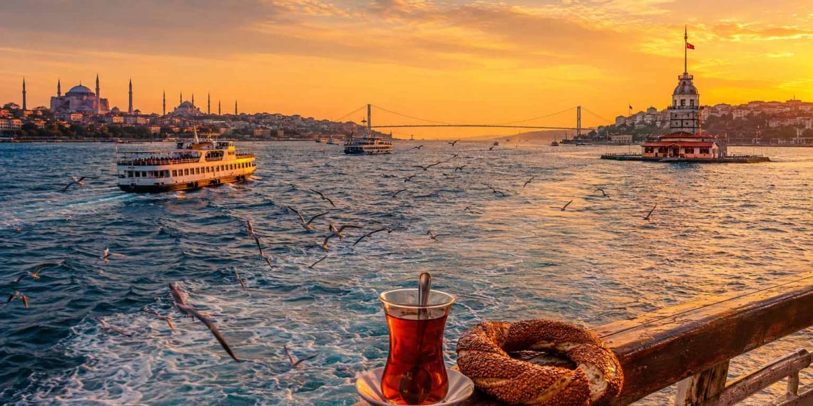 Un ferry municipal navigue sur le Bosphore à Istanbul au coucher du soleil, avec un verre de thé turc et un simit au premier plan, reliant la rive européenne historique à la rive asiatique authentique.