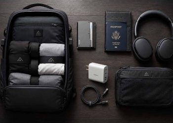 Les accessoires de voyage que j'utilise vraiment (Guide complet 2026) 2 Flat lay artistique des meilleurs accessoires de voyage minimalistes et techniques de 2026 disposés sur une table en bois sombre.