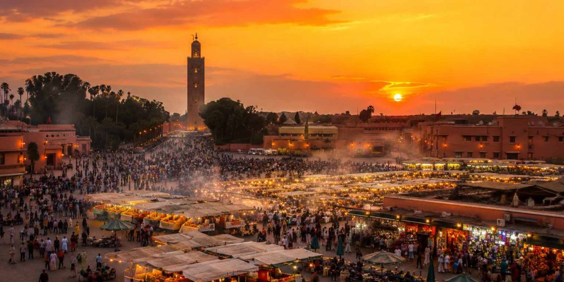 Ce que personne ne raconte sur un voyage à Marrakech 1 Vue panoramique de la place Jemaa el fna à Marrakech au coucher du soleil avec la fumée des stands de nourriture et le minaret de la Koutoubia.
